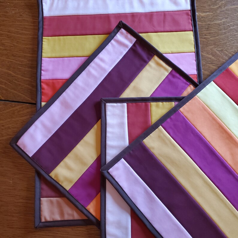 May include: Three quilted placemats with a striped pattern in shades of purple, pink, yellow, and orange. The placemats have a brown border.