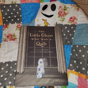 May include: A children's book titled "The Little Ghost Who Was a Quilt" with a cover illustration of a ghost wearing a quilt. The book is on a colorful patchwork quilt with floral and polka dot patterns. The book's author is Riel Nason.