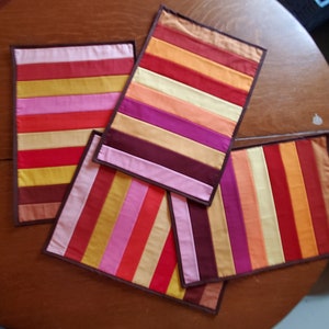 May include: Four colorful striped placemats with brown trim. The placemats are made of fabric and have a variety of colors, including red, orange, yellow, pink, purple, and brown.