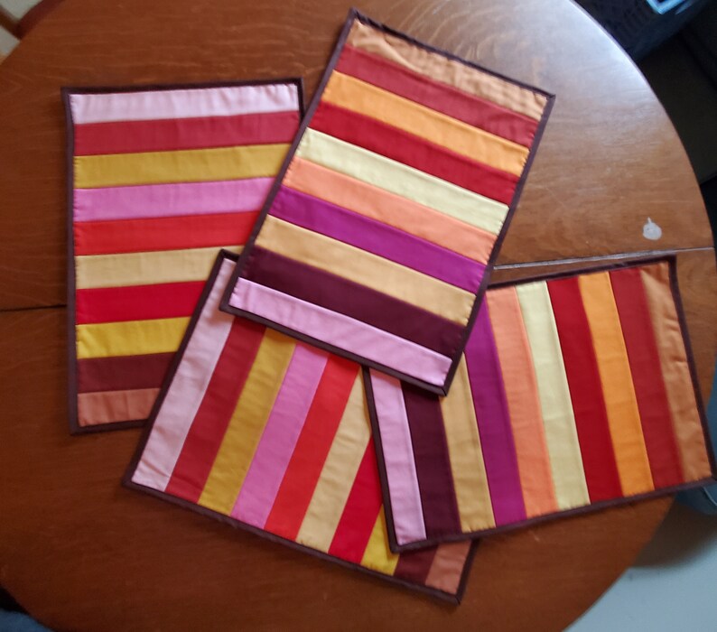 May include: Four colorful striped placemats with brown trim. The placemats are made of fabric and have a variety of colors, including red, orange, yellow, pink, purple, and brown.