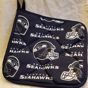 Pode incluir: Um pegador de panela acolchoado azul com o logotipo do Seattle Seahawks repetido por toda parte. O logotipo apresenta um capacete de futebol branco com um pássaro no topo.