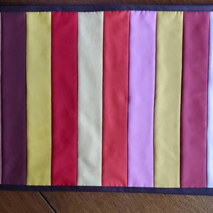May include: A long, rectangular table runner with a striped pattern. The stripes are in shades of brown, yellow, red, cream, pink, and white.