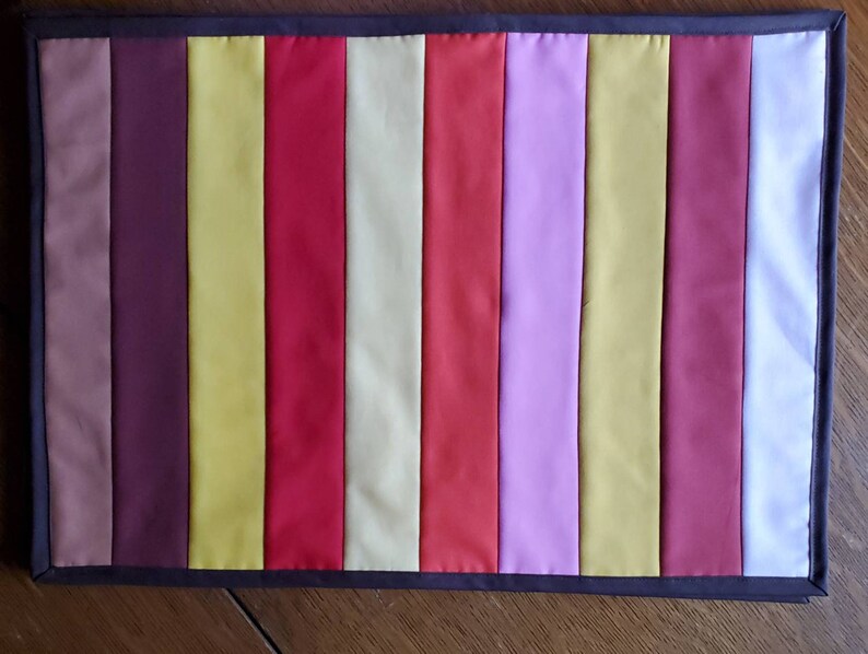 May include: A long, rectangular table runner with a striped pattern. The stripes are in shades of brown, yellow, red, cream, pink, and white.