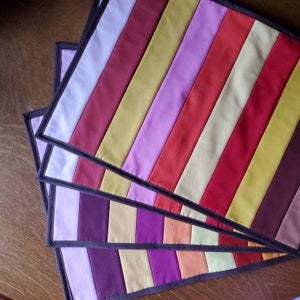 May include: Four quilted placemats with a striped pattern in shades of purple, red, yellow, and white. The placemats have a dark brown border.