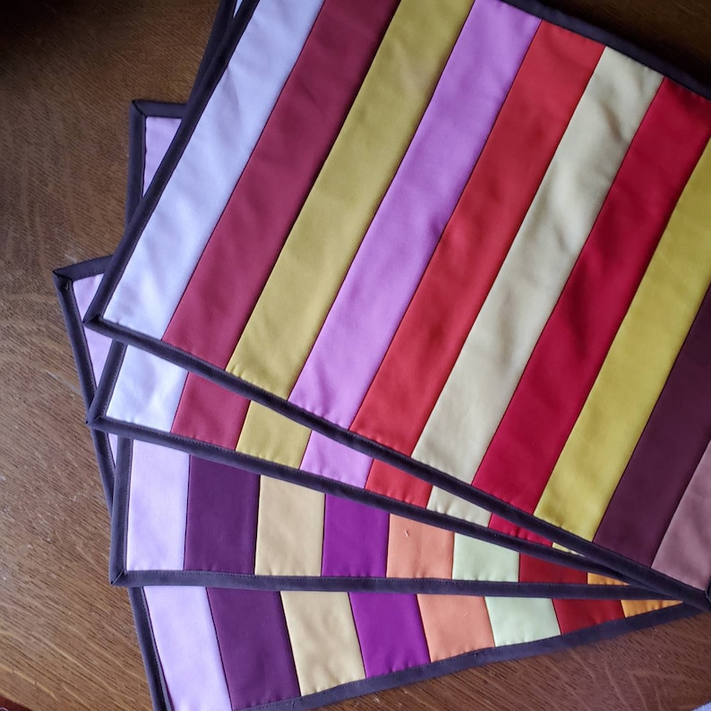 May include: Four quilted placemats with a striped pattern in shades of purple, red, yellow, and white. The placemats have a dark brown border.
