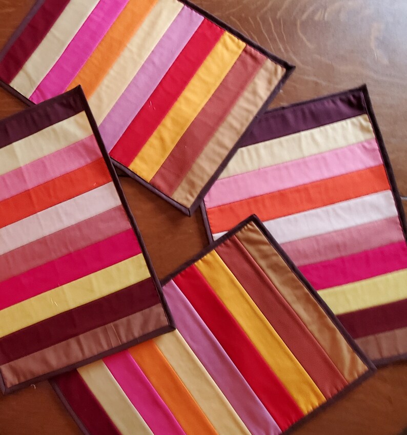 May include: Four colorful striped placemats with brown trim. The placemats are made of fabric and have a patchwork design.