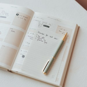 May include: An open planner with a green pen lying on the open page. The page has handwritten notes that say "Remember for next week: buy gifts, research trip, photo shoot pups".