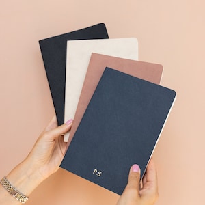 Peut inclure: Quatre cahiers empilés les uns sur les autres. Le cahier du dessus est bleu marine avec les lettres "P.S." en lettres dorées. Le deuxième cahier est de couleur brun clair. Le troisième cahier est de couleur crème. Le cahier du bas est noir.