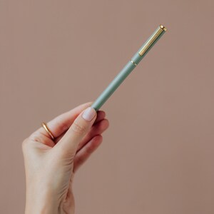 May include: A sage green ballpoint pen featuring a gold-toned clip and a matching gold-toned band at the center. The pen has a smooth, matte finish and a slender, cylindrical body design, providing a clean and functional writing instrument.
