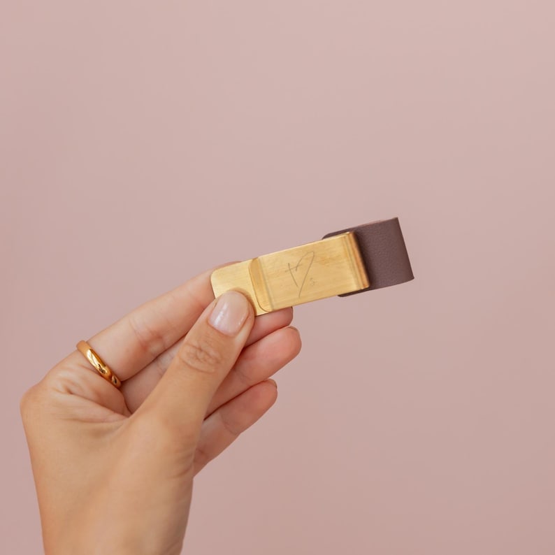 May include: A gold-colored money clip with a brown leather strap. The clip has an engraved design and is held by a hand with a gold ring. The background is a solid, soft pink.