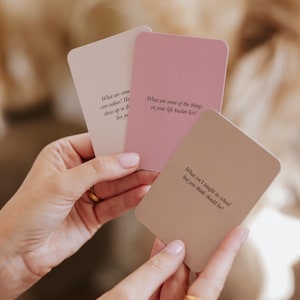 May include: Three rectangular cards in shades of pink and beige are held in a person's hands. Each card has a different question printed on it in black text. The questions are about core values, bucket lists, and what should be taught in school.