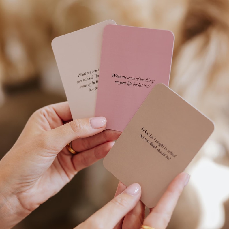 May include: Three rectangular cards in shades of pink and beige are held in a person's hands. Each card has a different question printed on it in black text. The questions are about core values, bucket lists, and what should be taught in school.