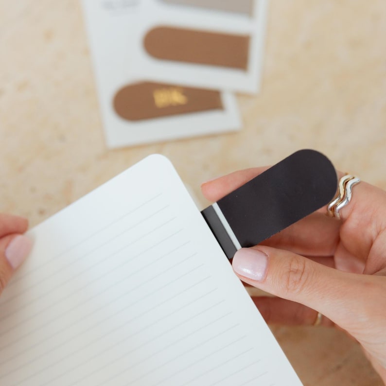 May include: A black and white bookmark is inserted into a notebook with lined pages. Additional brown and white bookmarks are visible in the background. The bookmark is held by a person with pink nail polish.