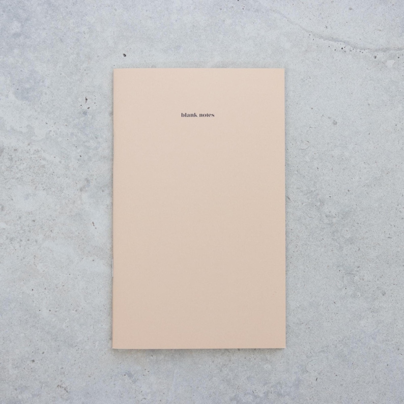 May include: A rectangular notebook with a solid light beige cover. The words blank notes are printed in small, black, lowercase sans-serif font near the top center of the cover. The notebook has a clean, flat, and uniform appearance.