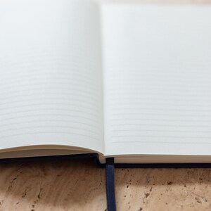 May include: Open notebook with lined pages and a dark blue ribbon bookmark. The pages are white and the cover is a dark blue. The notebook is open on a light brown surface.