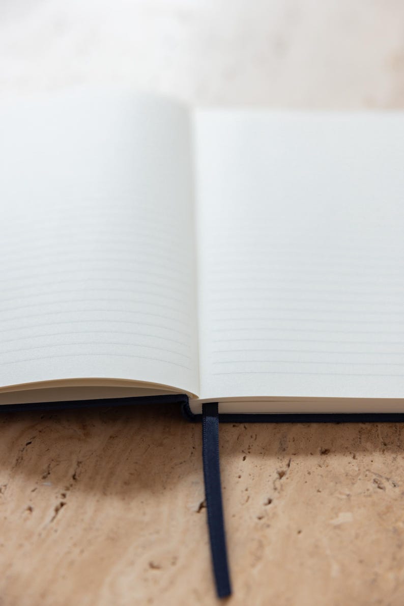 May include: Open notebook with lined pages and a dark blue ribbon bookmark. The pages are white and the cover is a dark blue. The notebook is open on a light brown surface.