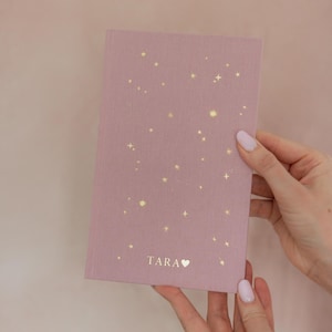 May include: A mauve-colored notebook with a constellation design in gold. The notebook has the name "TARA" printed at the bottom with a heart symbol. The notebook is held by a person.