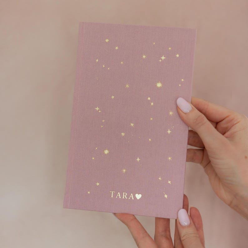 May include: A mauve-colored notebook with a constellation design in gold. The notebook has the name "TARA" printed at the bottom with a heart symbol. The notebook is held by a person.