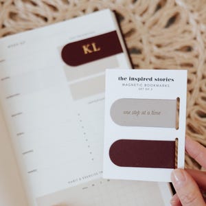 May include: A set of two magnetic bookmarks with the text "one step at a time". One bookmark is burgundy, the other is light gray. The bookmarks are on a white card with the text "the inspired stories MAGNETIC BOOKMARKS SET OF 2". A planner is in the background.