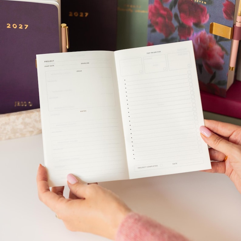 May include: An open planner with white pages featuring a project layout on the left side with sections for ideas and notes, and a top priorities checklist on the right side. The planner is held open, revealing printed text and lines for organization.