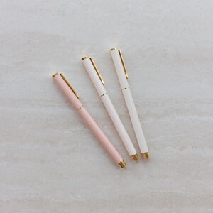 May include: Three slim ballpoint pens arranged diagonally. One pen features a soft pastel pink barrel, while the other two pens have white barrels. Each pen is accented with polished gold-tone metal clips, tips, and center rings for a refined look.