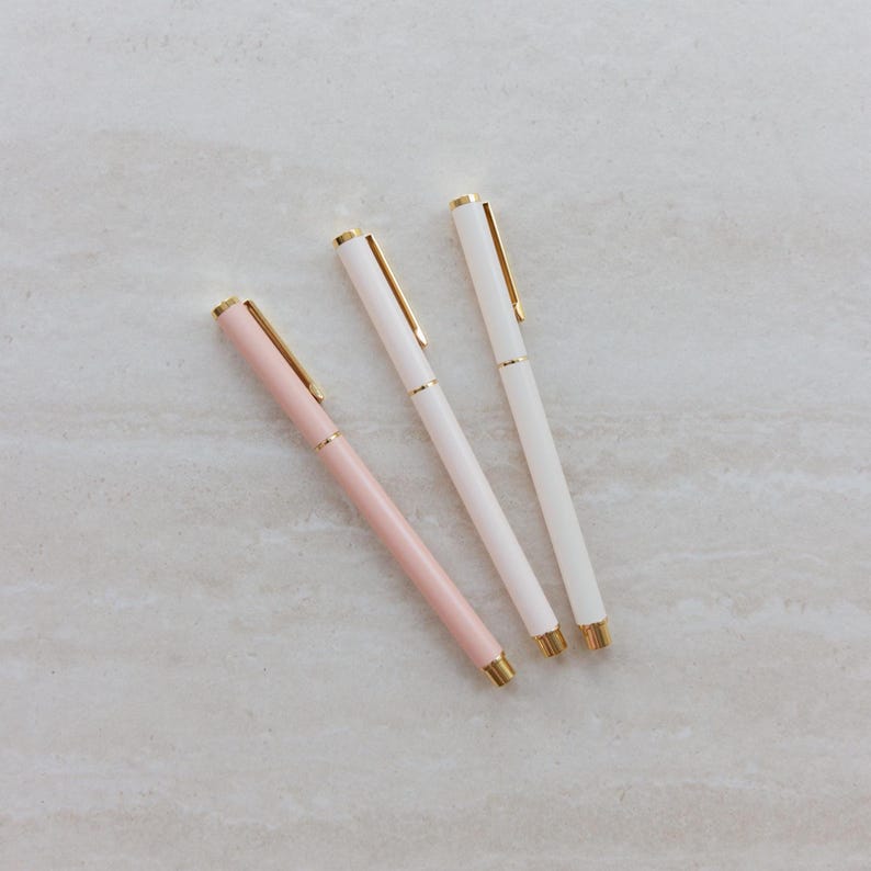 May include: Three slim ballpoint pens arranged diagonally. One pen features a soft pastel pink barrel, while the other two pens have white barrels. Each pen is accented with polished gold-tone metal clips, tips, and center rings for a refined look.