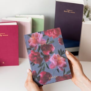 May include: A rectangular planner with a dark blue cover featuring a pink and red floral pattern, gold text reading "TWENTY TWENTY SEVEN" and "SARAH". Several other planners in solid colours like magenta, light pink, green, and dark purple are visible.