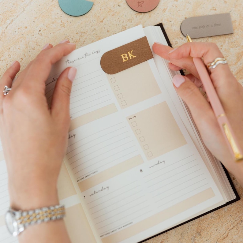 May include: A planner with a brown leather bookmark featuring the gold letters "BK". The open pages display a weekly layout with checkboxes and the text "These are the days". A pink pen rests on the right page.