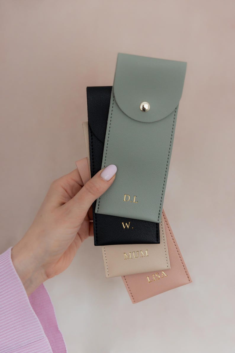 K&ouml;nnte beinhalten: Eine Sammlung von vier rechteckigen Etuis in Salbeigr&uuml;n, Schwarz, Beige und Altrosa. Jedes Etui hat einen goldenen Druckknopfverschluss und eine goldene Schriftpr&auml;gung. Das salbeigr&uuml;ne Etui ist mit D.L., das schwarze mit W., das beige mit MUM und das rosa mit LISA beschriftet.