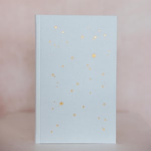 May include: A light blue journal with a soft cover. The cover is decorated with gold foil star shapes of varying sizes. The journal is closed and has a clean, minimalist design, perfect for writing or sketching.