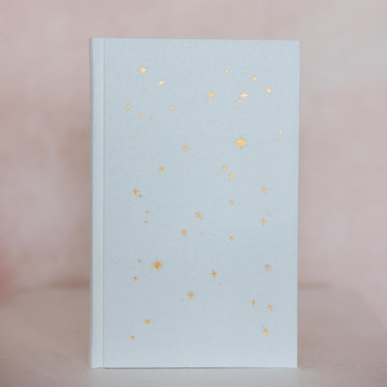May include: A light blue journal with a soft cover. The cover is decorated with gold foil star shapes of varying sizes. The journal is closed and has a clean, minimalist design, perfect for writing or sketching.