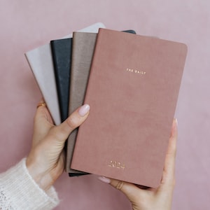 May include: A selection of five hardback journals in shades of dusty rose, grey, black, and brown. The front journal is dusty rose with the gold text "THE DAILY" and "2024". The journals are held in a person's hands against a pink backdrop.