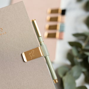 May include: A sage green pen with gold accents is held by a gold-colored pen holder attached to a beige notebook. The pen holder has a leather strap and a stylized initial design. Other notebooks and accessories are in the background.