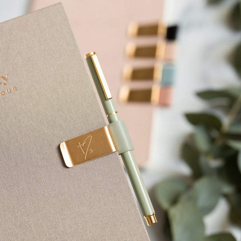 May include: A sage green pen with gold accents is held by a gold-colored pen holder attached to a beige notebook. The pen holder has a leather strap and a stylized initial design. Other notebooks and accessories are in the background.