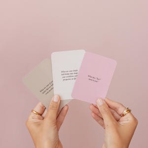 May include: Three pastel-colored cards with rounded corners are held in front of a pink background. The cards are beige, white, and pink, each with a question printed in black. The questions are about beliefs, habits, and the meaning of love.