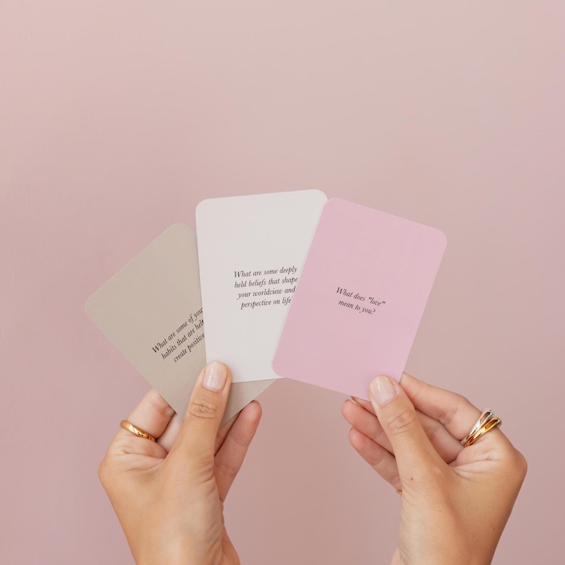 May include: Three pastel-colored cards with rounded corners are held in front of a pink background. The cards are beige, white, and pink, each with a question printed in black. The questions are about beliefs, habits, and the meaning of love.