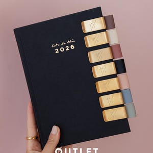 May include: A black 2026 planner with the text "let's do this 2026" in gold lettering on the cover. The planner features seven gold-toned metal page markers with a heart-shaped logo, each attached to a coloured tab in shades of brown, cream, and blue.