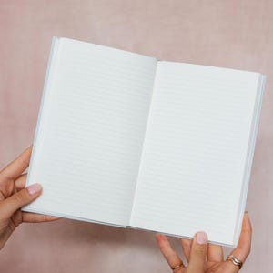 May include: An open, lined notebook with a light blue cover. The pages are white with horizontal lines. The notebook is held open, displaying the blank pages. The notebook is a simple design, perfect for writing or sketching.
