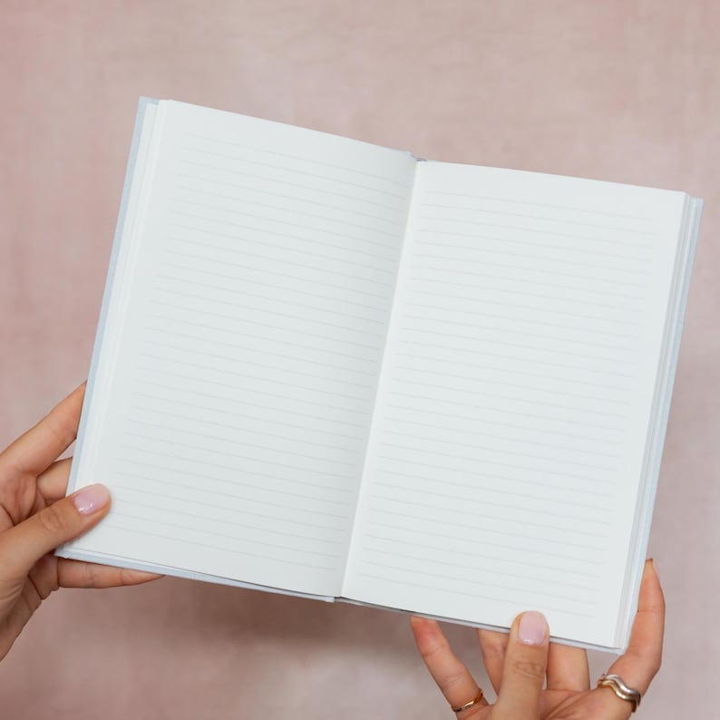 May include: An open, lined notebook with a light blue cover. The pages are white with horizontal lines. The notebook is held open, displaying the blank pages. The notebook is a simple design, perfect for writing or sketching.