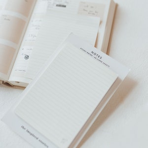 May include: A white notepad with lined pages and a white cover. The cover has the text "NOTES" and "STICKY NOTES - 60 LINED SHEETS" printed in black. The notepad is sitting on top of a closed book.