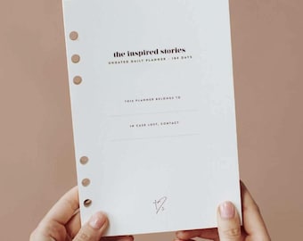 Könnte beinhalten: Ein weißer Tagesplaner mit dem Text "the inspired stories" und "UNDATED DAILY PLANNER - 180 DAYS". Der Planer hat einen Bereich für "THIS PLANNER BELONGS TO" und "IN CASE LOST, CONTACT".