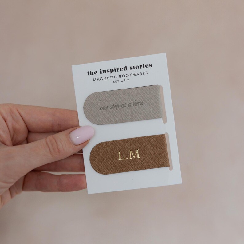 May include: Two magnetic bookmarks on a white card. The top bookmark is light gray with the text "one step at a time." The bottom bookmark is brown with the initials "L.M." in gold. The card reads "the inspired stories MAGNETIC BOOKMARKS SET OF 2."