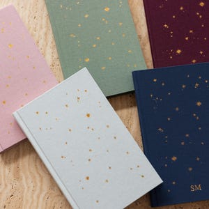 May include: Five hardcover journals in various colors: light pink, sage green, burgundy, light blue, and navy. Each journal features a constellation design in gold foil. The navy journal has the initials "SM" in gold foil.