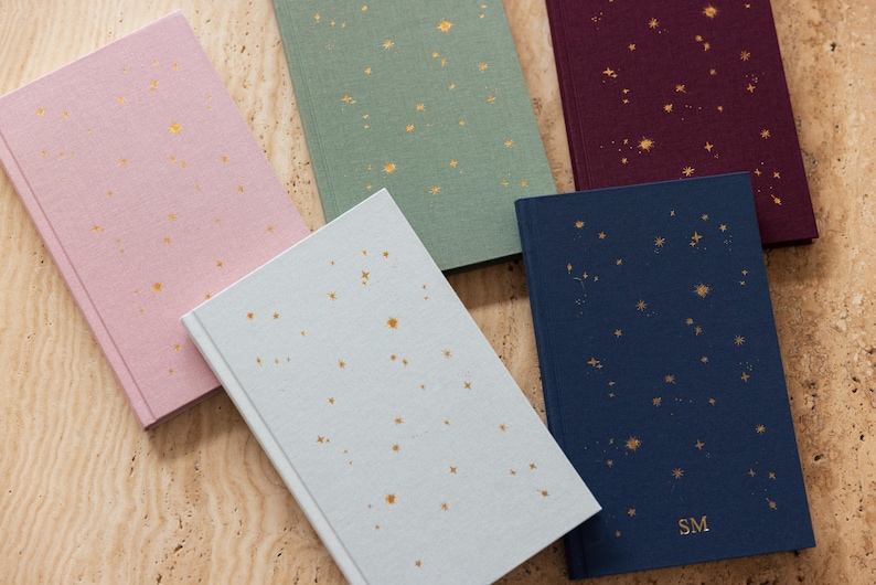 May include: Five hardcover journals in various colors: light pink, sage green, burgundy, light blue, and navy. Each journal features a constellation design in gold foil. The navy journal has the initials "SM" in gold foil.