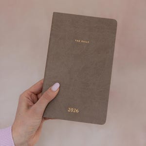K&ouml;nnte beinhalten: Ein rechteckiger Planer mit einem strukturierten taupefarbenen Einband. Auf der Vorderseite befindet sich der goldgepr&auml;gte Text THE DAILY im oberen Bereich und das Jahr 2026 in Gold am unteren Rand. Der Planer hat abgerundete Ecken und eine matte Oberfl&auml;che.