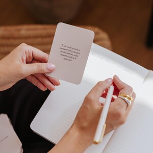 May include: A person holding a card with text and writing in a notebook with a pen. The card reads: "If you could go back to a moment from your past for five minutes, what moment would it be and why?"