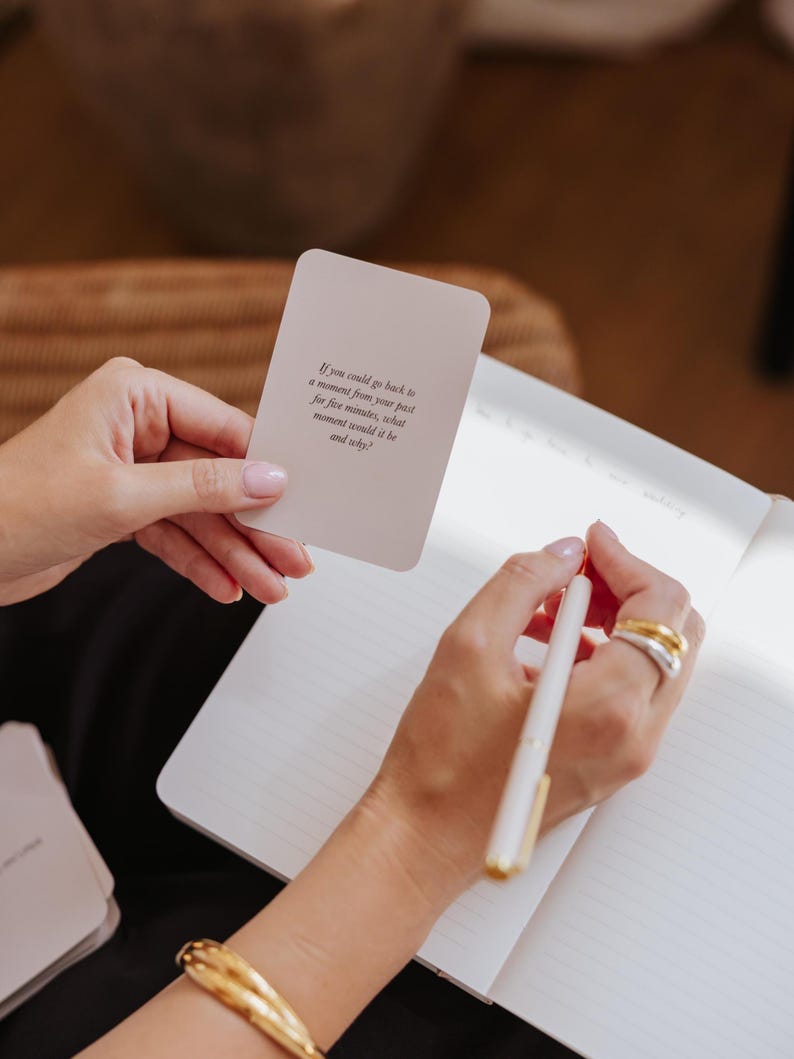 May include: A person holding a card with text and writing in a notebook with a pen. The card reads: "If you could go back to a moment from your past for five minutes, what moment would it be and why?"