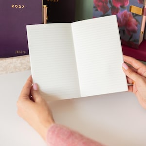 May include: An open notebook with white lined pages held in place. In the background, there is a dark purple notebook with the text 2027 printed in gold, and another notebook featuring a floral pattern with pink and red flowers on a dark background.
