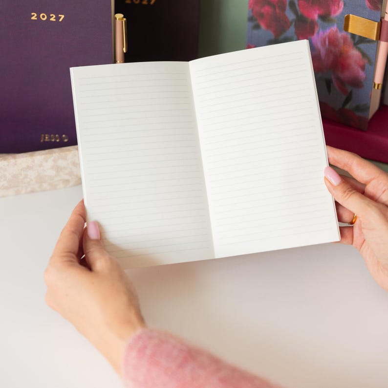 May include: An open notebook with white lined pages held in place. In the background, there is a dark purple notebook with the text 2027 printed in gold, and another notebook featuring a floral pattern with pink and red flowers on a dark background.