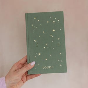 May include: A green hardcover journal with a celestial design of gold stars. The word "LOUISE" is printed in a simple font at the bottom of the cover. The journal is held by a person.